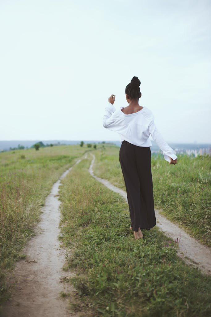 A woman walks barefoot on a grassy rural path, embracing nature and solitude.